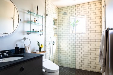 vanity with undermount sink and black vanity top, round mirror, glass shelves, white toilet, walk-in shower with glass door, subway tile wall, black hexagon shower tile floor