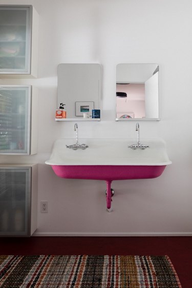 pink cast iron bathroom sink with double faucets, two rectangular mirrors, three vertical storage units, multicolored area rug