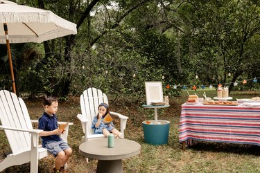 Two small kids sitting in white Adirondack chairs under fringed umbrella enjoying potato chips