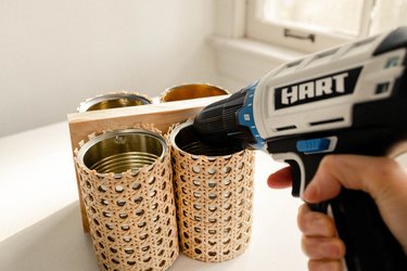 Attaching empty soup cans wrapped in cane webbing to wood board with HART power drill