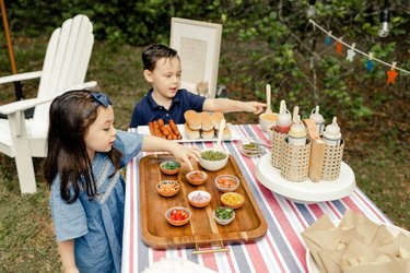 Two small kids pointing to toppings on the hot dog bar table