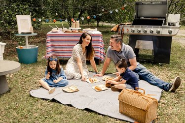 Parents and two small kids sitting on picnic blanket enjoying hot dogs and chips