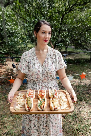 Woman holding wooden tray with six gourmet hot dogs and potato chips