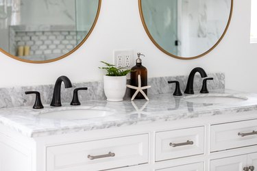 white vanity with gray marble countertop, double sinks with black faucet, vase with green plant, starfish decoration, soap dispenser, two round mirrors with gold trim