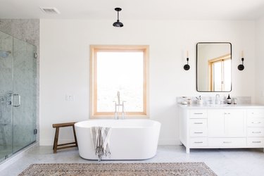 large bathroom with freestanding white tub, glass shower with large marble tile, rectangular window, black pendant light fixture, white vanity with marble top, rectangular mirror with black trim, two black sconce light fixtures, brown area rug