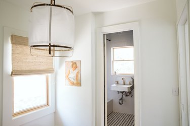 white ceramic wall-mounted sink with gold faucet, black and white hexagon tile floor, hanging pendant light fixture, window with tan Roman shade