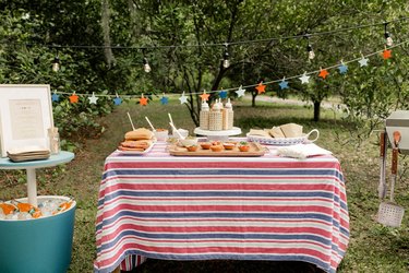 Gourmet hot dog bar with red, white and blue star garland, striped tablecloth and beverage cooler table with framed menu