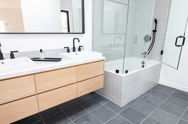 light wood floating vanity with two ceramic vessel sinks, black faucets, rectangular mirror, gray square tile floor, white tub with glass shower doors