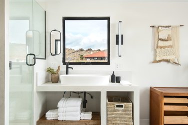 white stone bathroom vanity, white ceramic vessel sink with black faucet, open window, hanging tapestry, storage basket, white towels, wood storage unit, glass shower door