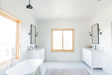 freestanding white tub with silver faucet, double vanities on opposite sides of the bathroom, matching rectangular mirrors with black trim, two windows, multicolored area rug, black pendant light fixture