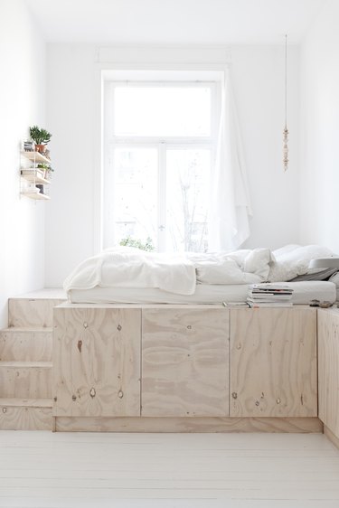 studio bedroom idea with a tall bed built out of plywood with storage underneat and stairs to the left. White bedding is on top.
