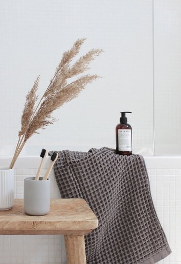 spa bathroom ideas in all white bathroom with natural wooden stool next to bathtub and gray waffle cotton towel