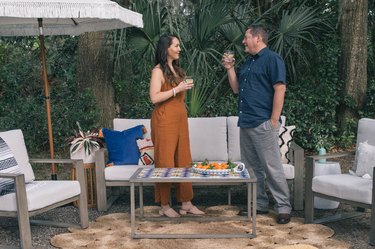 Woman and man standing in outdoor patio with DIY tiled coffee table, colorful pillows and fringe umbrella