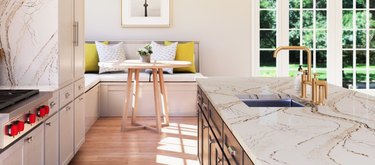 Gold-veined quartz countertop colors, light wood floors, brass faucet, breakfast nook, round wood dining table, yellow and white cushions.