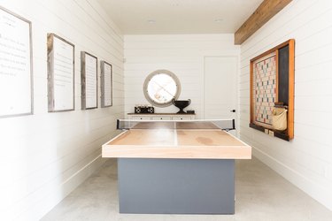 attic game room with light wood and gray ping pong table, concrete floors, white walls, round window.