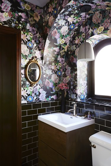 Black floral wallpaper in maximalist bathroom with wood vanity and arched window