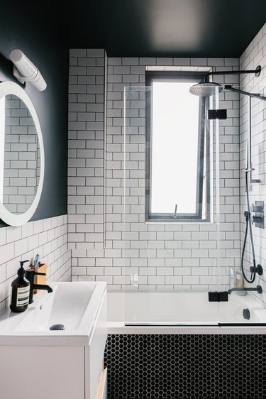 small white sink, round mirror with overhead lighting, black hexagon tile on the bathtub, white subway tile on the wall, deep bathtub with a black faucet and showerhead