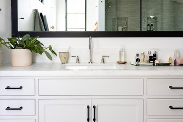 gray bathroom vanity cabinet, undermount sink, silver faucet and handles, rectangular mirror with black trim, vase with green plant, mirrored tray with various bathroom products