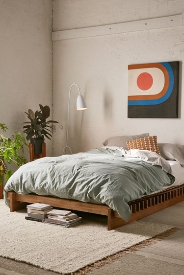 Concrete floors and walls in light color, natural wood platform bed with light gray bedding, white reading lamp, plants, contemporary art print, beige area rug. Basement Bedroom Ideas
