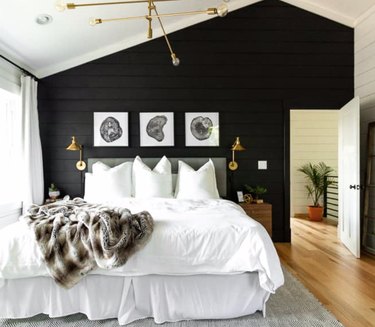 Black paneled accent wall, white bedding, gray headboard, brass modern chandelier, light wood floors, brass sconces, black and white art above bed, faux fur throw with rustic colors