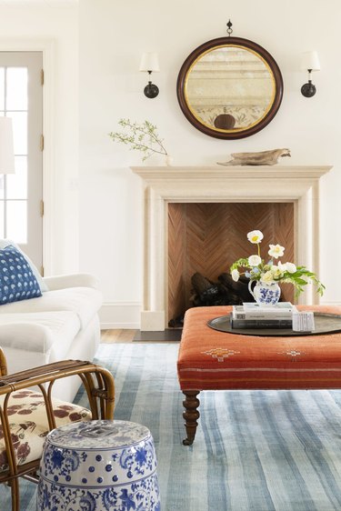 white living room with blue rug, orange ottoman and traditional fireplace with mirror on top