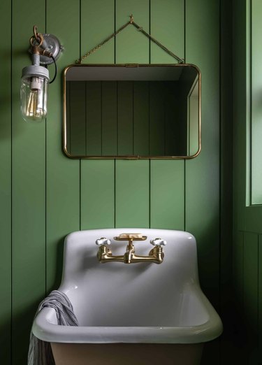 Moss green shiplap walls, vintage mirror, sconce, sink, brass faucet in bathroom with rustic colors