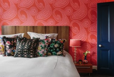 vermillion color wallpaper, wood headboard, floral throw pillows, navy blue door, lamp with red patterned lampshade.