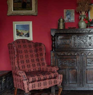vermillion color patterned high back chair, dark red wall, dark wood credenza, art on walls.
