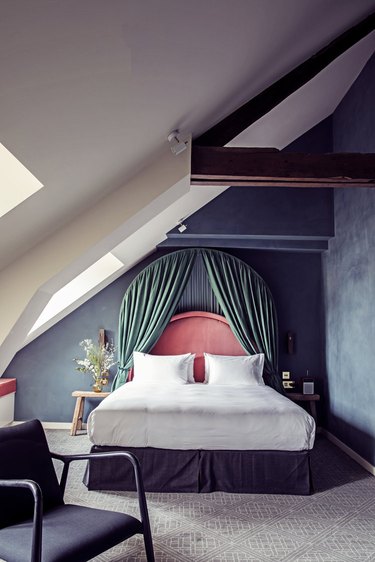 Bedroom with blue walls that appear lime washed and a bed with green velvet curtains over a salmon headboard and dark purple bedskirt.