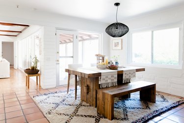 A black pendant light over dark wood dining furniture on a diamond pattern rug, and terra-cotta tile floor.