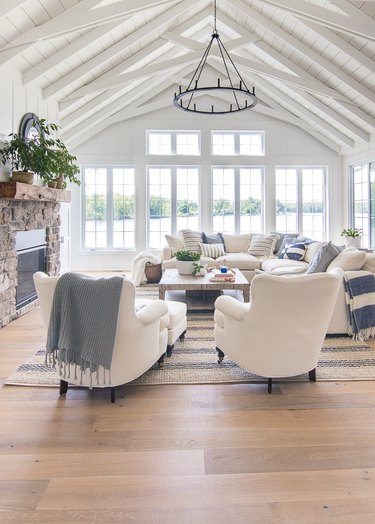 coastal living room white arched ceiling with white beams, iron chandelier, stone fireplace, light oak floors, white wing back chairs, white sectional.