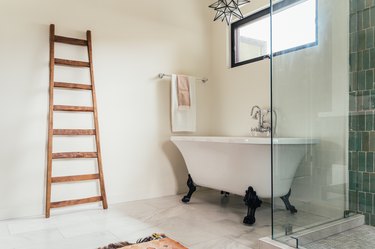 clawfoot tub with black angled legs, decorative wood ladder leaning on the wall, glass shower door with green vertical tiles