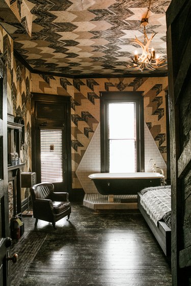 black clawfoot tub on a platform in a bedroom, patterned walls and ceiling, dark wood floors, an armchair and a bed are also seen in the room