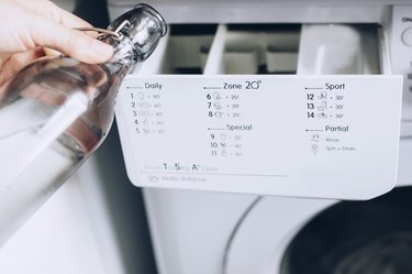 Hand about to pour soap mixture from glass bottle into detergent compartment of white washing machine