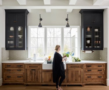 black kitchen cabinet idea with rustic wooden lower cabinets with white countertops and a white farmhouse sink. Above the counter on either side of a large window are two black cabinets with glass doors.
