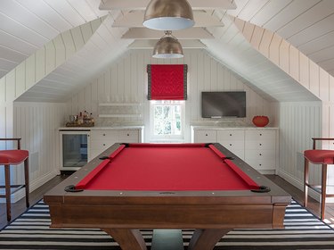 attic game room with red pool table, red stools, red window shade, white shiplap walls and ceiling, metal pendant lamps, black and white striped rugs, flat screen, built-in cabinets.