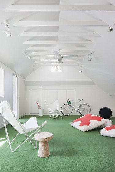 White attic game room with green astro turf, white canvas butterfly chairs, orange and white floor cushions, bicycle, end table, ceiling fan.