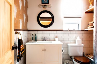 bathroom with small white vanity, undermount sink, round mirror with black trim, subway tile wall, built-in shelving, toilet with wood lid