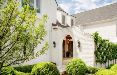 painting exterior brick on 0ff-white painted brick house, arched entry, plants, tree, hedge.