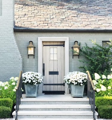 painting exterior brick with pale green on home with faded wood door, matching outdoor sconces, flowered plants in pots.