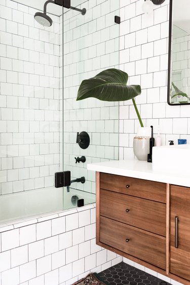 white subway tile on walls and side of bathtub, glass shower door, black showerhead, faucet and handles, wood floating vanity, black hexagon floor tile