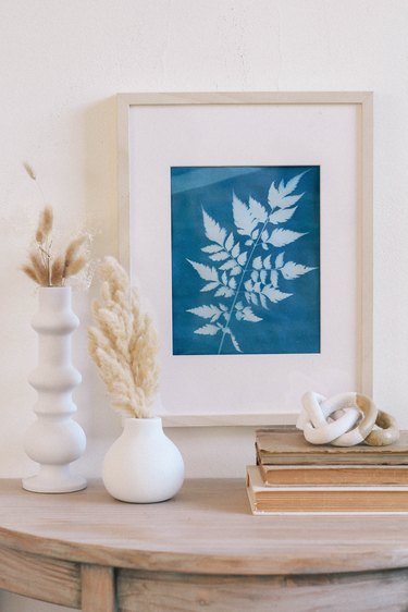 Framed blue sun print hung on wall above table with white vases, dried flowers, books and a ceramic knot