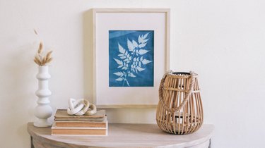 Framed blue sun print hung on wall above table with white vases, dried flowers, books and a rattan candle holder