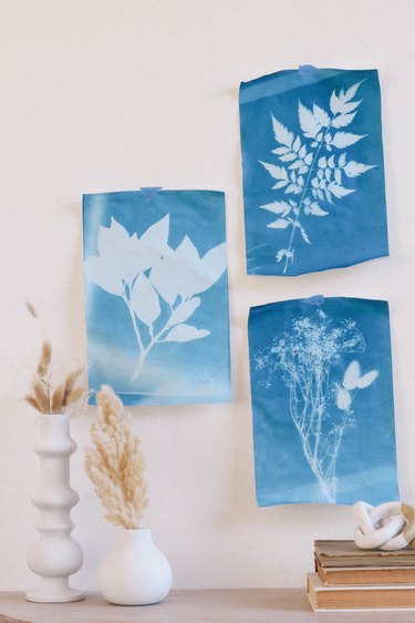 Three blue sun prints hung on wall with tape above table with white vases and books