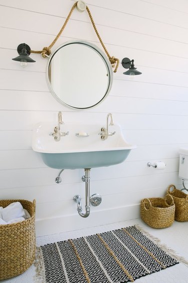 coastal bathroom with white and light blue trough sink, round nautical mirror, black finish dock lights, white shiplap walls.
