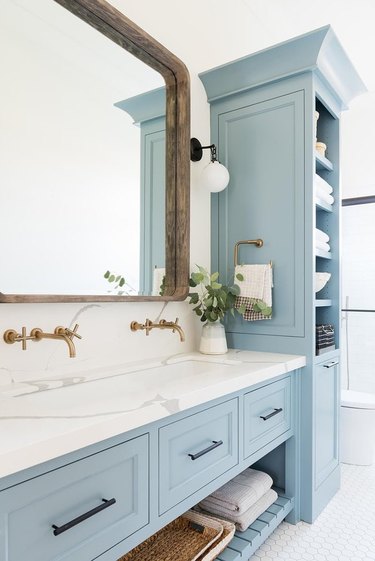 coastal bathroom with large farmhouse mirror, marble vanity, brass faucets, blue cabinets and shelves.