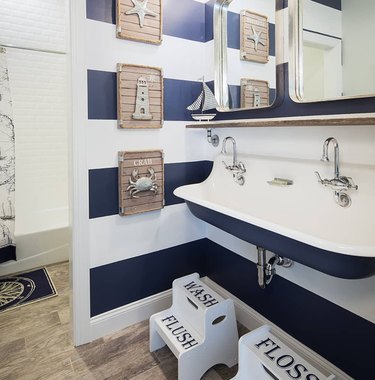 coastal bathroom with blue and white striped walls, trough sink, nautical themed artwork, foot stools.