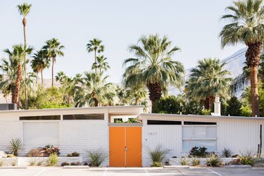 white home exterior on midcentury Palm Springs house with orange door surrounded by palm trees