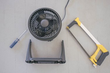 Black table fan with its stand removed next to screw driver and hacksaw