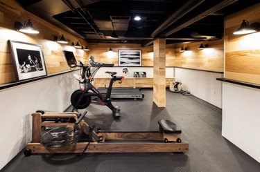 basement ideas with Gym equipment, rowing machine, bike, weights, padded flooring, black and white photos, light wood paneled and white walls.